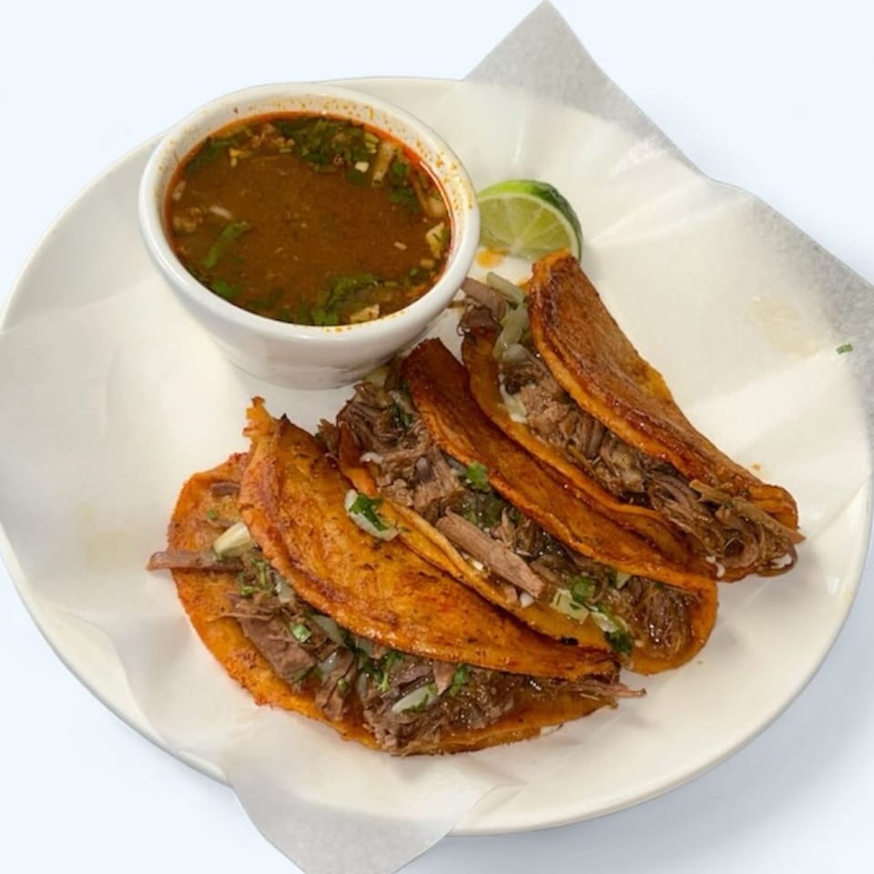 Three birria tacos with shredded beef, cilantro, and onions, served with consommé and a lime wedge.