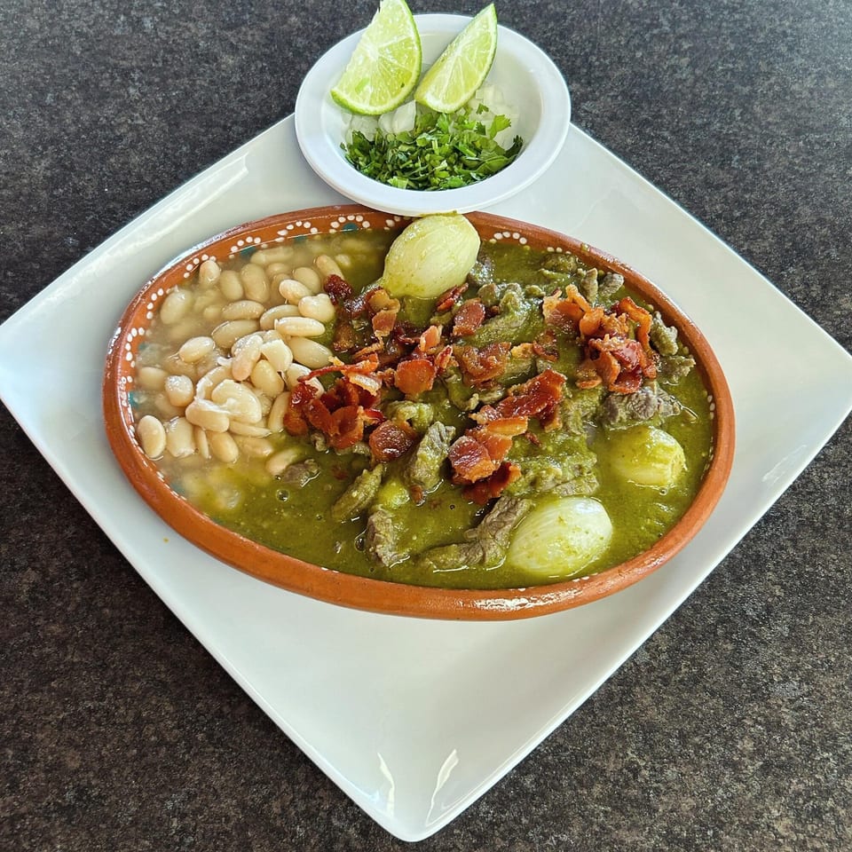 Mexican stew with green sauce, white beans, meat, bacon, and a side of lime and cilantro.